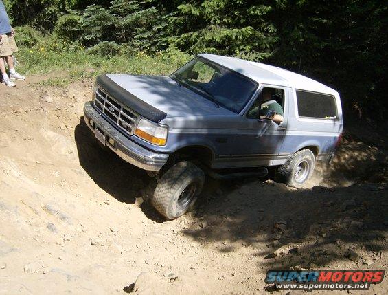 Ford Bronco driving on a dirt trail with trees in the background. Driver is visible