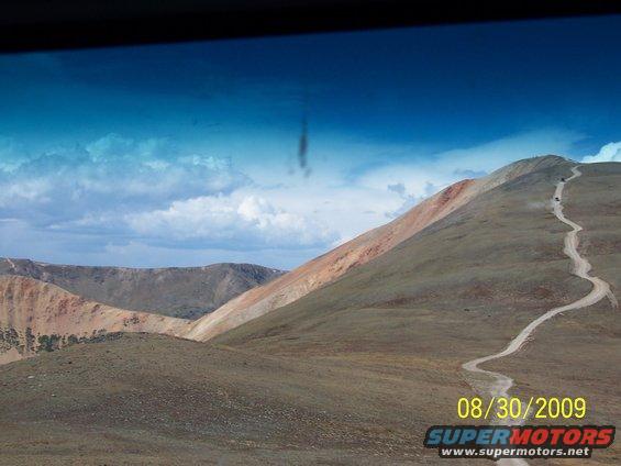 Several vehicles drive up a winding dirt road on a mountain under a cloudy blue sky