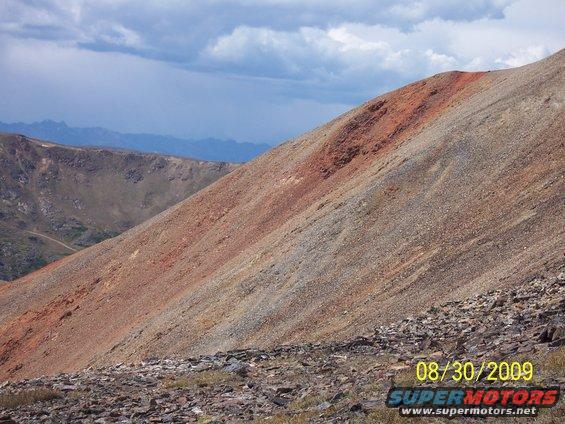 Mountain slope with red and gray rocks under a cloudy sky. A dirt road is visible in the distance