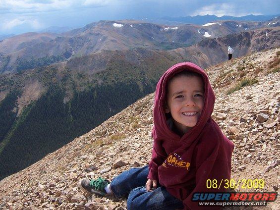 Child in maroon hoodie and jeans sits on a rocky mountain slope with a vast mountain range in the background