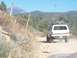 1990 Ford Bronco - A white 1990 Ford Bronco kicks up dust as it travels along a remote dirt road, with mountains and dry vegetation in the background.