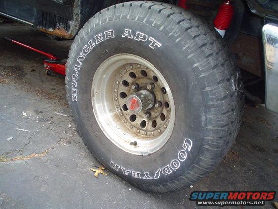 Close-up of a Goodyear Wrangler APT tire on a vehicle with a red jack underneath. Rust is visible on the vehicle's body