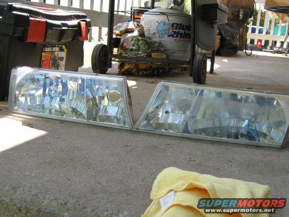 Two clear plastic car headlights with silver trim, one slightly fogged, lying on concrete. A yellow microfiber cloth is in the foreground