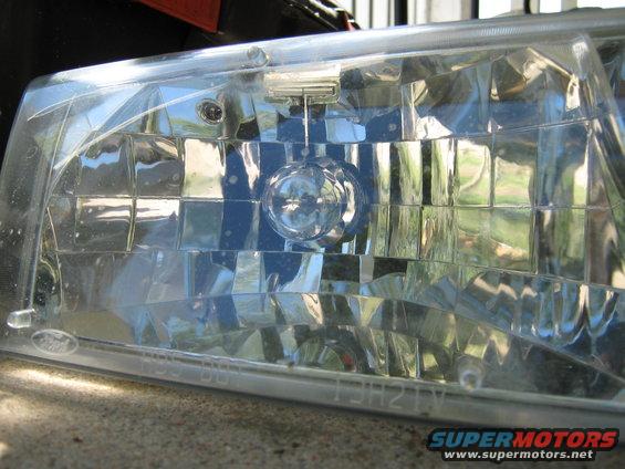 Close-up of a clear plastic headlight assembly with internal reflectors and bulbs. Text is visible on the bottom edge