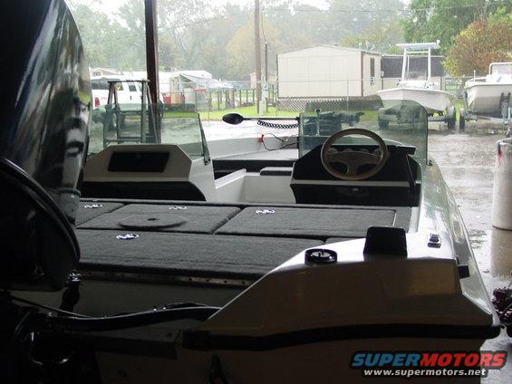 Boat interior with steering wheel, seats, and dashboard. Other boats and buildings in background