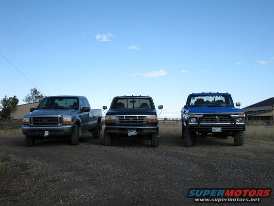 Three pickup trucks parked in a line on a dirt road. The truck on the left is blue, the middle truck is black, and the truck on the right is blue