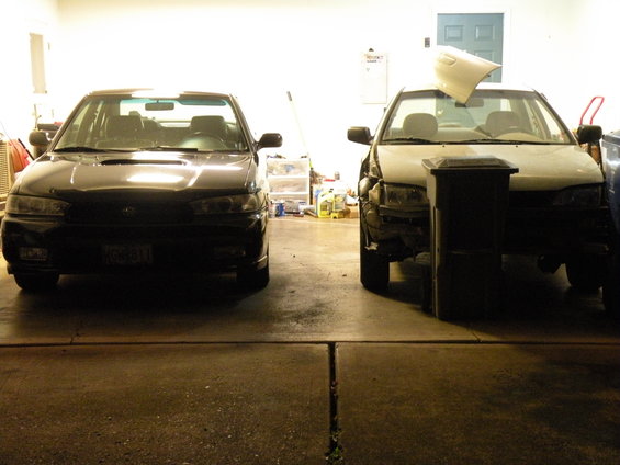 Black 1998 Subaru Legacy sedan parked next to a white Subaru sedan with front damage