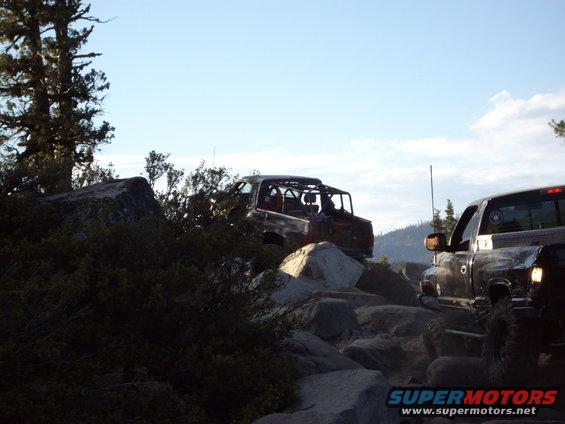 Two off-road vehicles navigate a rocky trail under a clear sky