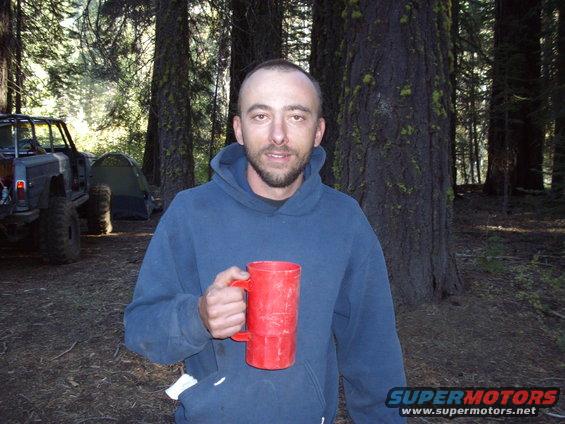 Man holding a red mug in a forest campsite with a modified off-road vehicle and tent in the background