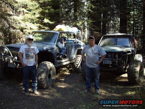 Two guys stand next to heavily modified off-road vehicles in a wooded area