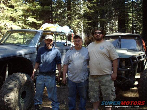 Three men stand in front of two modified off-road vehicles in a wooded area. The vehicle on the left has a large tire. The vehicle on the right has a roll cage