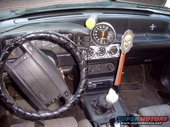 Car interior showing dashboard, steering wheel with Ford logo, aftermarket tachometer and gauges, and gear shifter