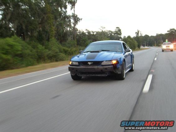 Blue Ford Mustang with black stripe and missing front bumper driving on highway, another car in distance