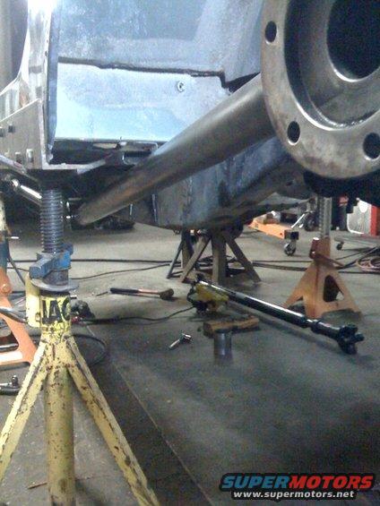 Vehicle frame and suspension parts on jack stands in a workshop. A driveshaft lies on the floor
