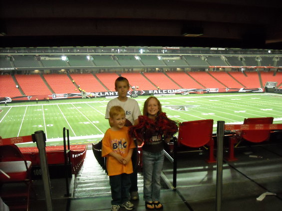 Three children stand in stadium seating with a football field in the background