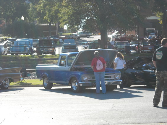 Blue and silver pickup truck with hood open, people looking at engine. Other vehicles in background