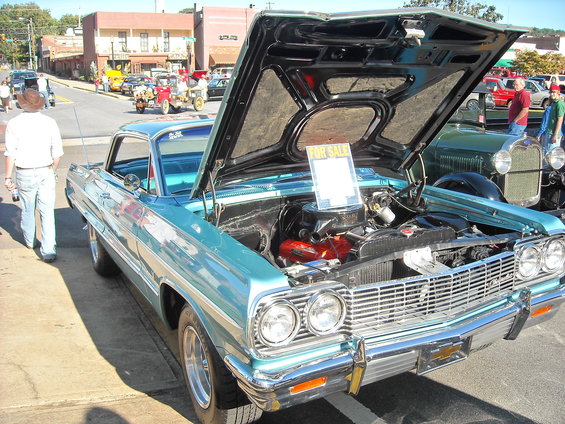 1964 Chevrolet Impala with hood open, "FOR SALE" sign on display, parked next to a green 1920s Ford Model A