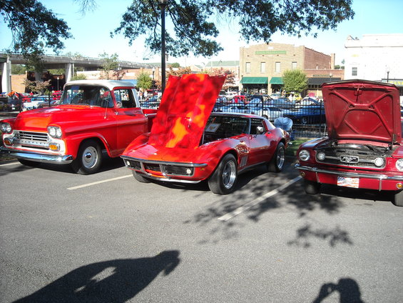 Three red classic cars parked in a lot: a 1950s pickup, a 1969 Corvette with hood open, and a Ford Mustang with hood open