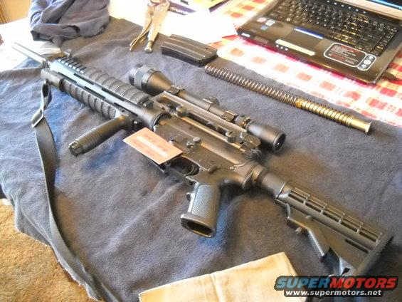 AR-15 style rifle with scope, magazine, and spring on a table next to a laptop