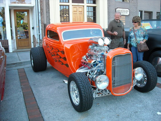 Orange hot rod with flame decals and a large supercharged engine. Two people stand nearby