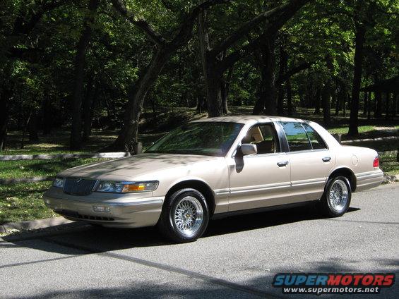 Tan sedan parked on a road with trees in the background. It has chrome wheels and a chrome grille