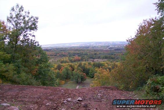 Autumn landscape viewed from a dirt embankment, with colorful trees and a distant town under a cloudy sky. Supermotors.net logo in lower right