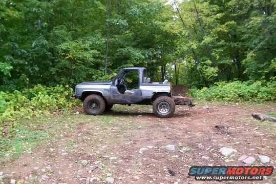 Off-road SUV with no doors parked in a wooded area. It has large tires and a trailer hitch