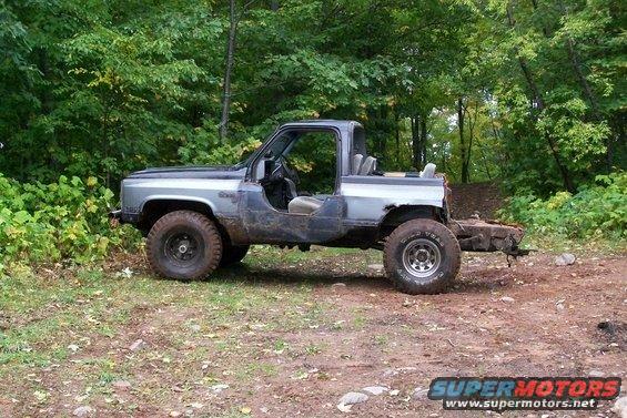 Modified SUV with removed doors and rear section, parked on a dirt path in a wooded area