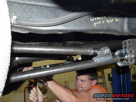 Mechanic works on the undercarriage of a vehicle, with yellow writing on the frame reading 'wrench pull = bar'