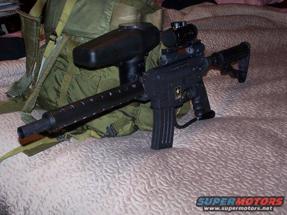 Paintball gun with scope and hopper next to a green backpack on a bed