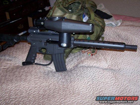 Black paintball gun with a large hopper and scope, resting on a bedspread