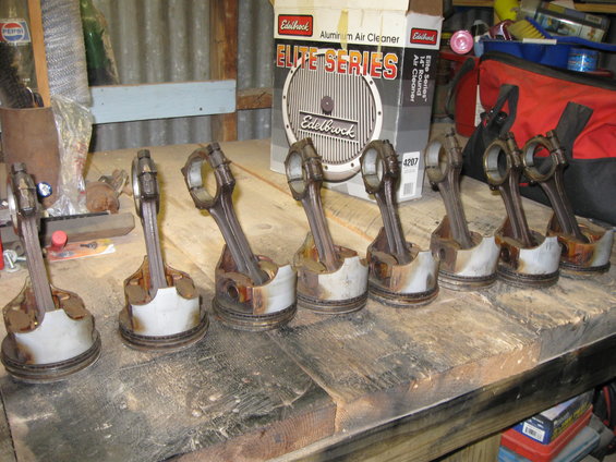 Eight used pistons with connecting rods arranged on a wooden workbench, with an Edelbrock air cleaner box in the background