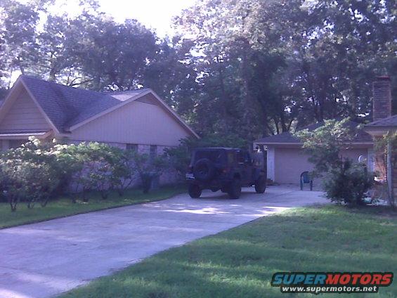 Black Jeep Wrangler parked in driveway, spare tire on rear door, trees and house in background