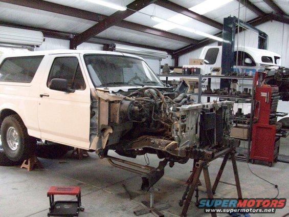 White SUV with front end removed, on jack stands in a workshop. Another white vehicle is visible in the background