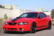 2004 Ford Mustang - A red 2004 Ford Mustang coupe is parked outside industrial buildings under a clear sky.