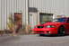 2004 Ford Mustang - A red 2004 Ford Mustang is parked in front of a warehouse-style building with overgrown weeds.