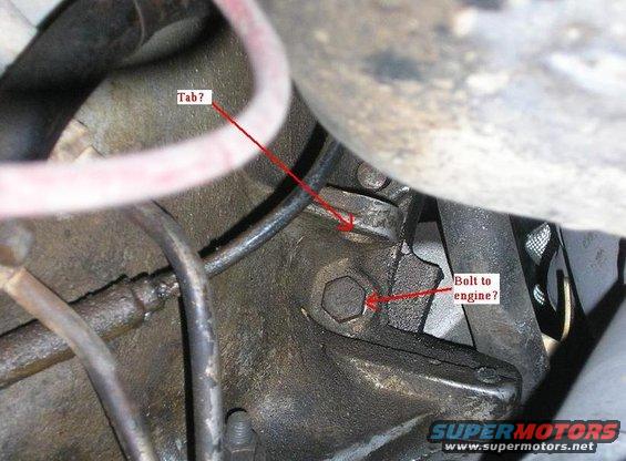 Close-up of engine component with red arrows pointing to a tab and a bolt. Hoses and wires are visible