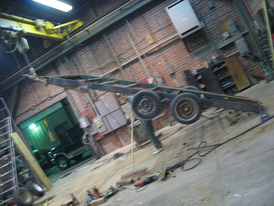 Trailer chassis with dual wheels in a workshop, with a dark SUV visible in the background