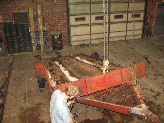 Man working on a red utility trailer suspended by a chain hoist in a garage