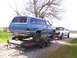 1900 Ford C-350 (Centurion Conversions) - A blue SUV is loaded onto a trailer, with a white pickup truck parked behind it on a gravel surface.