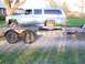 1900 Ford C-350 (Centurion Conversions) - A light blue SUV is loaded onto a trailer, parked on a gravel surface with grass and a fence in the background.