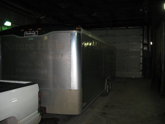 Silver enclosed trailer with 'Nature Hills Nursery' text, parked next to a white pickup truck in a garage