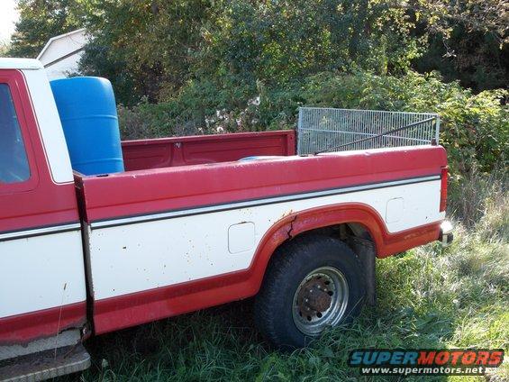 Side view of a red and white pickup truck bed with a blue barrel and metal rack inside. Some rust and damage visible