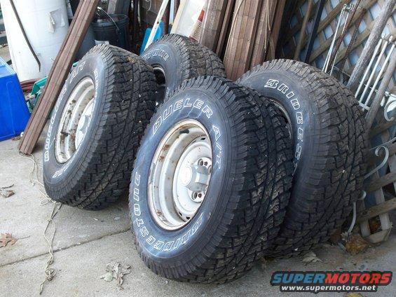Four Bridgestone Dueler A/T tires on steel wheels, leaning against a fence