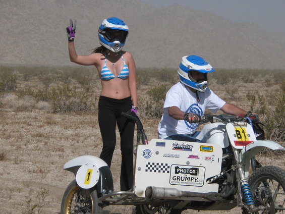 Person in bikini and helmet gives peace sign next to motorcycle with sidecar