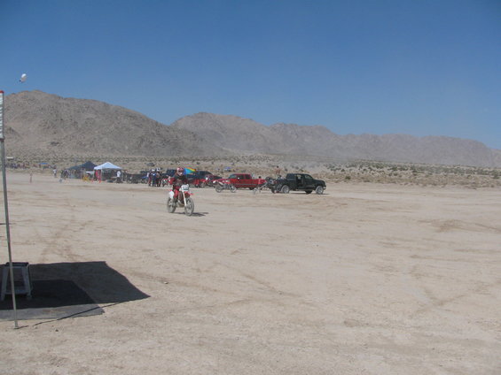 Dirt bike rider in a desert landscape with mountains in the background and several parked vehicles
