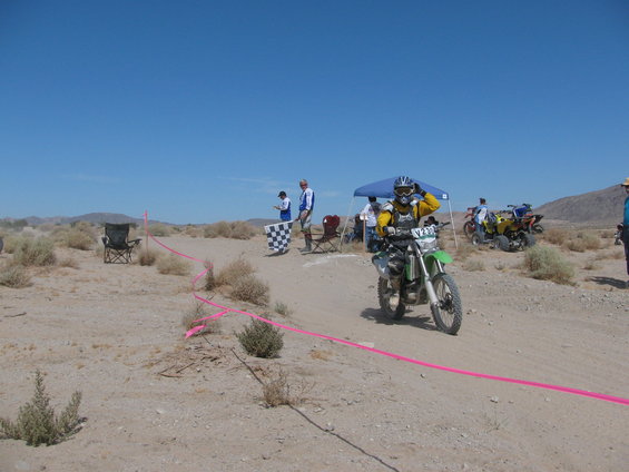 Dirt bike rider approaches finish line with checkered flag and officials. Other ATVs and bikes in background