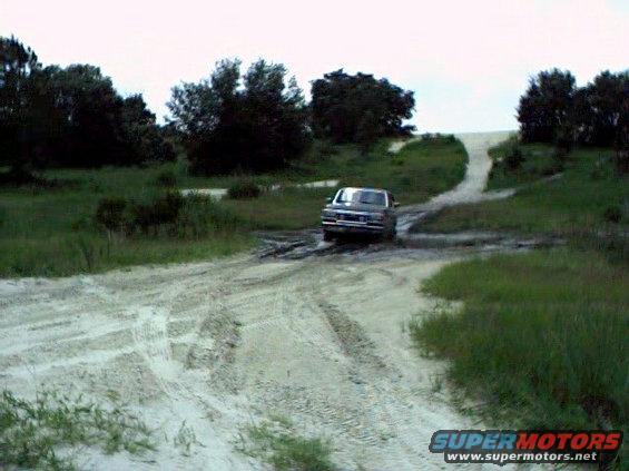 Gray Ford pickup truck driving through mud on a dirt road with trees and a hill in the background