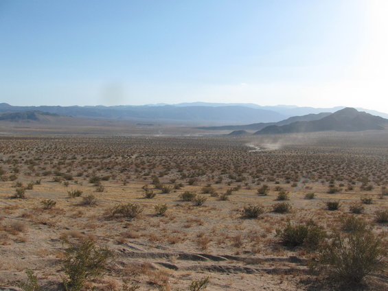 Vast desert landscape with sparse bushes and distant mountains under a clear blue sky. Dust clouds are visible in the distance