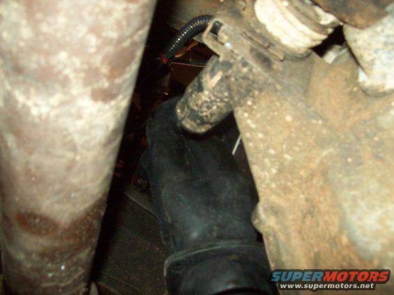Undercarriage view showing exhaust pipe and suspension parts. A gloved hand is visible near a wire harness